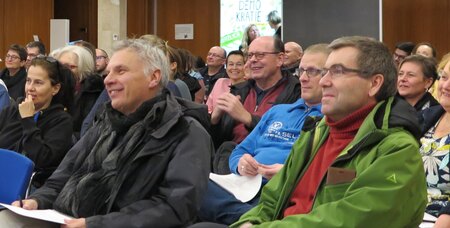 Viele Menschen sitzen auf Stühlen in einem Saal. Sie hören jemandem zu und lachen.